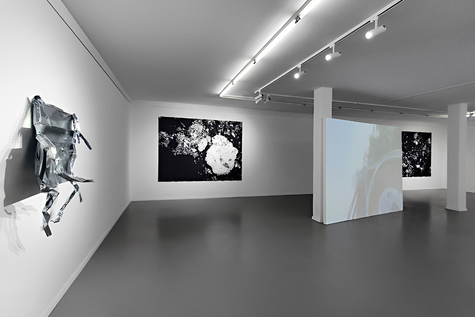 Exhibition view of “Is History OK?” by Johannes Hugo Stoll, showing a gallery space with a projection screen, wall-mounted works, and white structural columns.