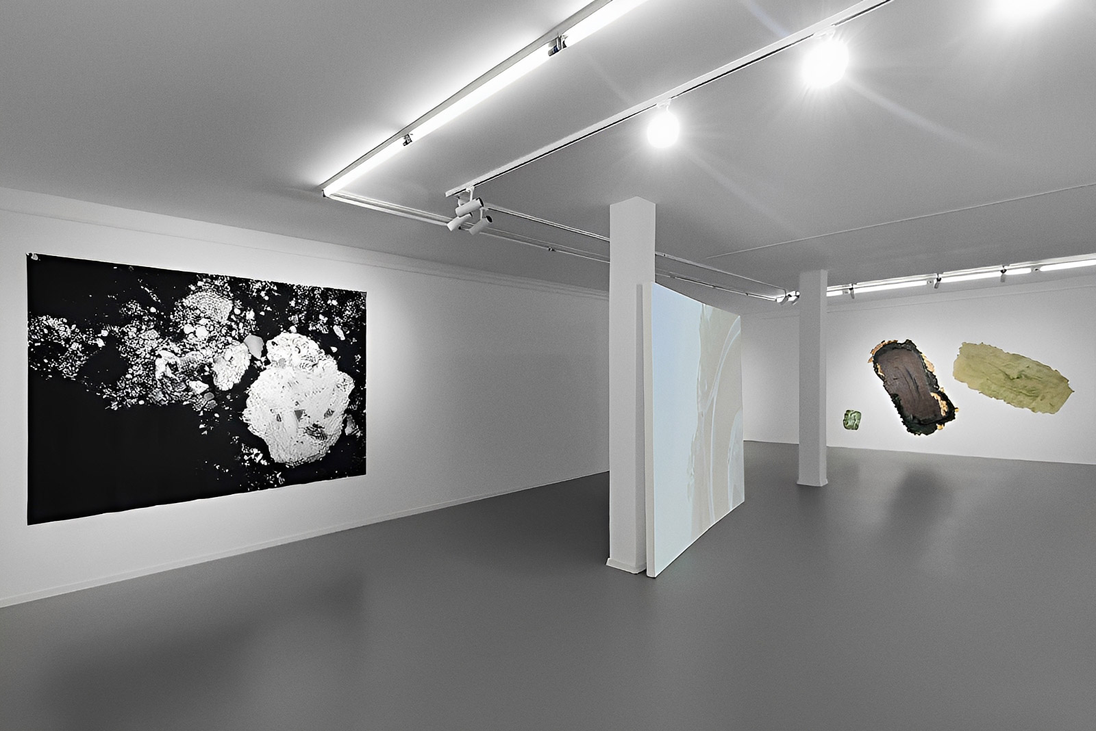 Exhibition view of “Is History OK?” by Johannes Hugo Stoll, showing a large black-and-white wall-mounted work, a central projection screen between white columns, and additional abstract works on the far wall.