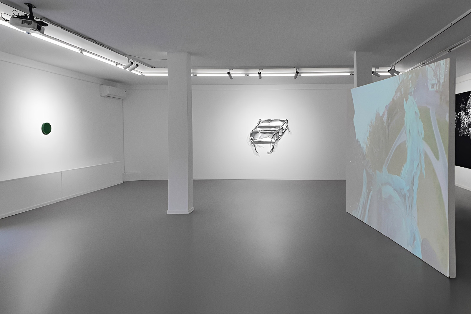 Exhibition view of “Is History OK?” by Johannes Hugo Stoll, showing a large projection screen in the foreground, a suspended metallic work on the back wall, and a small green circular work on the left wall.