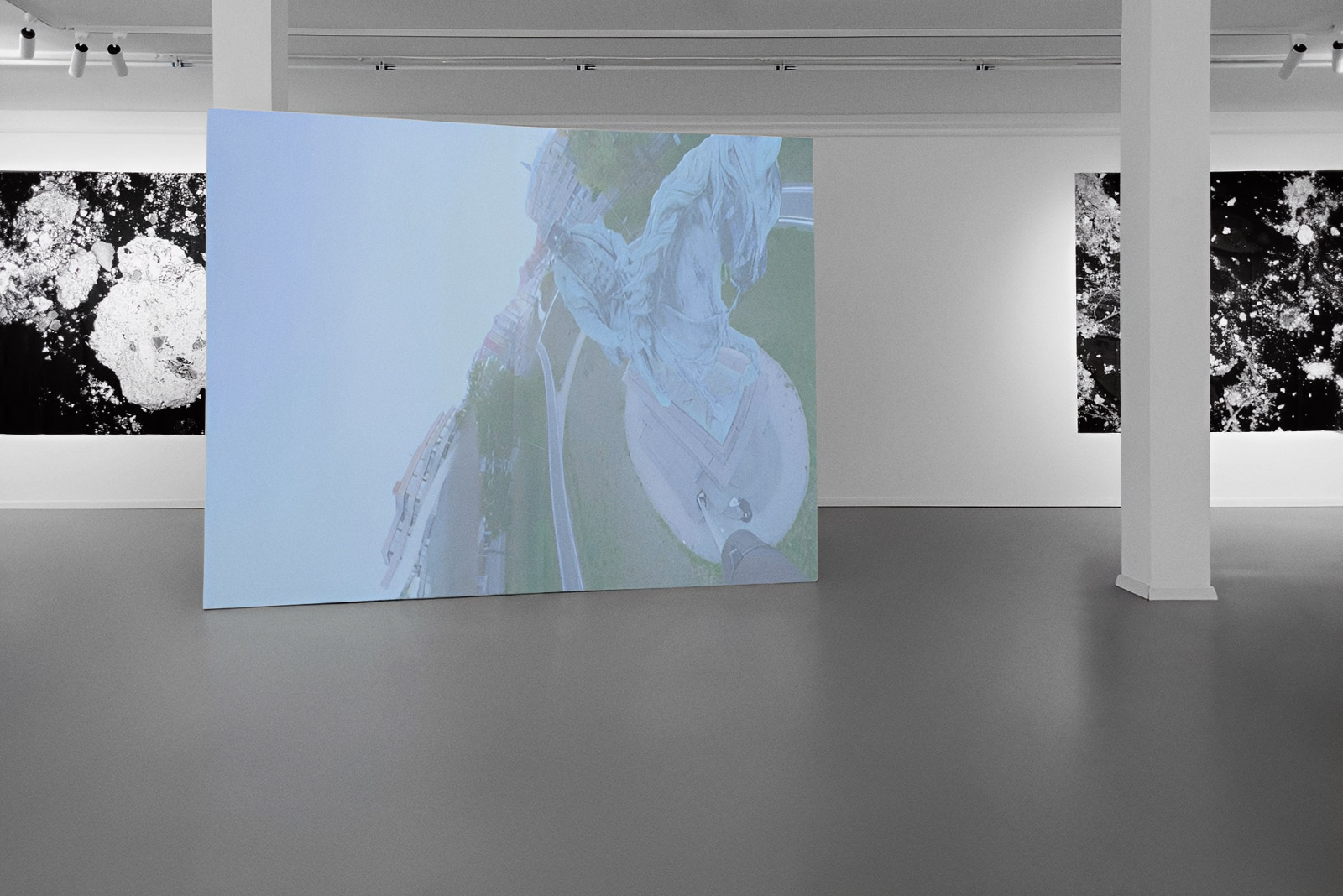 Exhibition view of “Is History OK?” by Johannes Hugo Stoll, showing a large freestanding projection screen in the foreground, framed by white columns and black-and-white works on the surrounding walls.