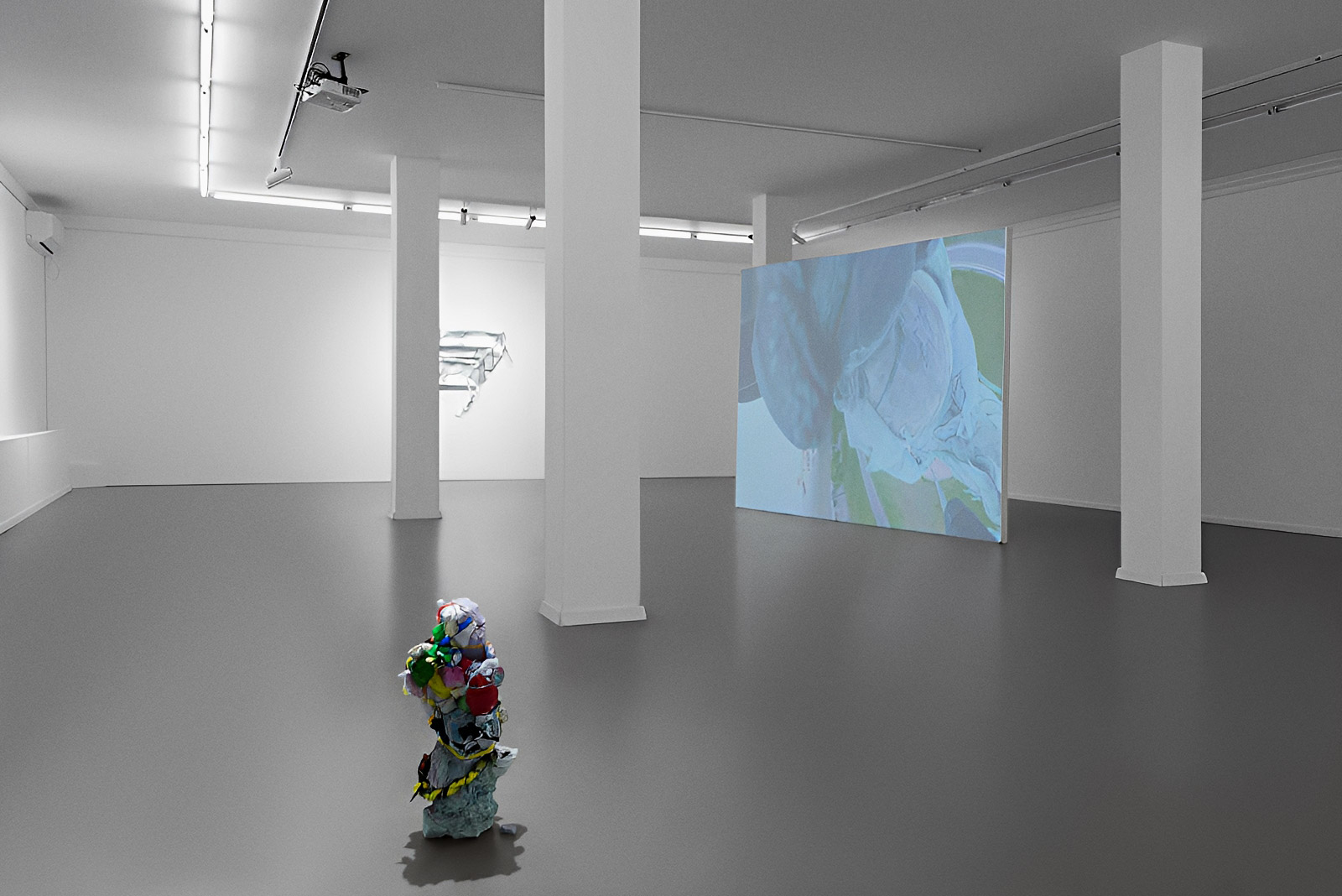 Exhibition view of “Is History OK?” by Johannes Hugo Stoll, showing a small sculptural work in the foreground, a freestanding projection screen between white columns, and a suspended metallic work in the background.